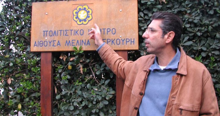 A man is shown from the waist up, pointing to a sign with Greek lettering.