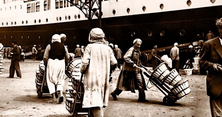 A black and white photo of men transporting goods in a shipping yard. A cargo ship is in the background.