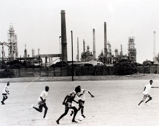 Black and white photo of five people playing soccer. In the background, the soccer field is bordered by a fence; beyond the fence are multiple industrial smokestacks.