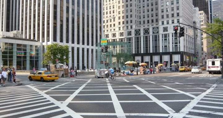 A photo of a crosswalk in New York City taken from a street corner. Vendors, pedestrians, and taxis line the street and sidewalk.