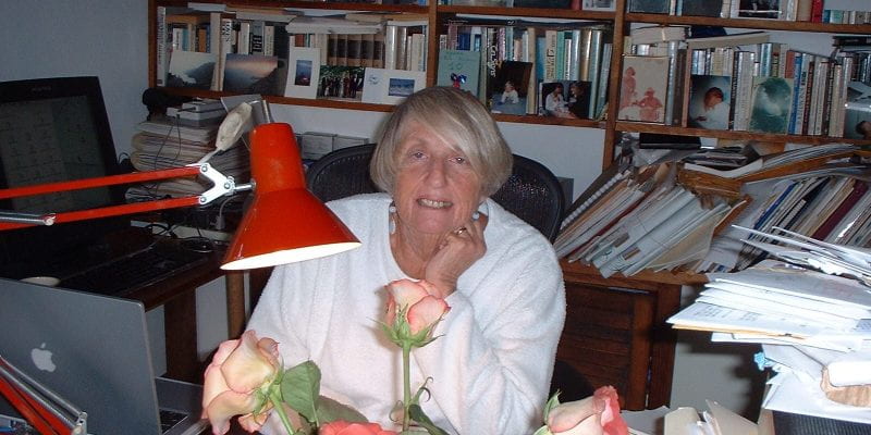 A photo of Helene Moglen sitting at her desk, resting her chin on one hand. A vase of flowers and a red reading lamp are in the foreground of the photo; a bookshelf filled with books is behind Helene.