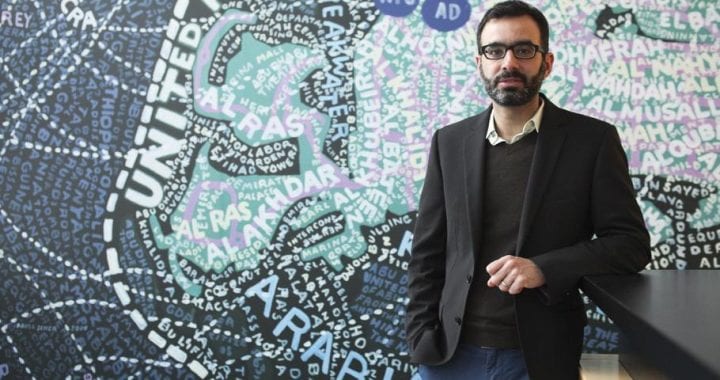 A photo of Ahmed Kanna standing in front of a mosaic of a map of the Arab gulf.